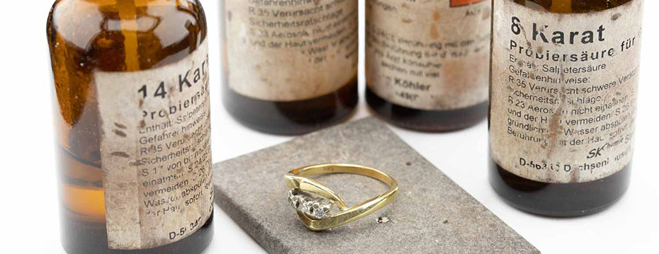 Ein goldener Ring neben verschiedenen braunen Fläschen mit Probiersäure.